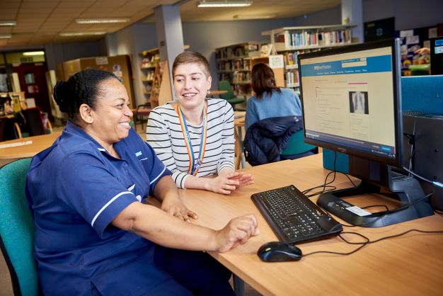 Two female colleagues sitting at a computer looking at the screen. One is a nurse wearing nurse scrubs and the other is wearing casual clothes. She is helping the nurse use the computer.