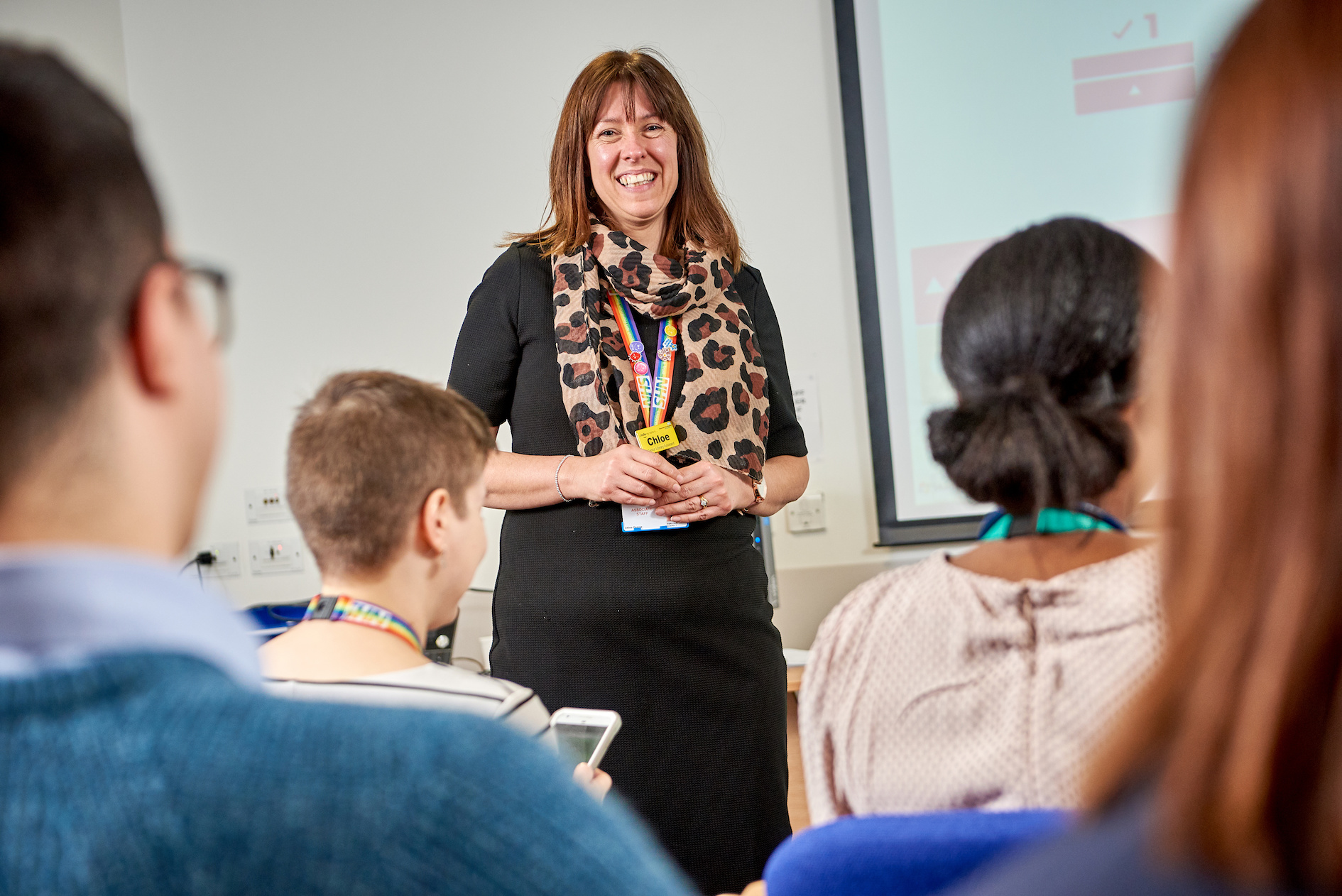 A female library professional is standing in front of her colleagues leading a meeting. Her colleagues are sitting down facing her.