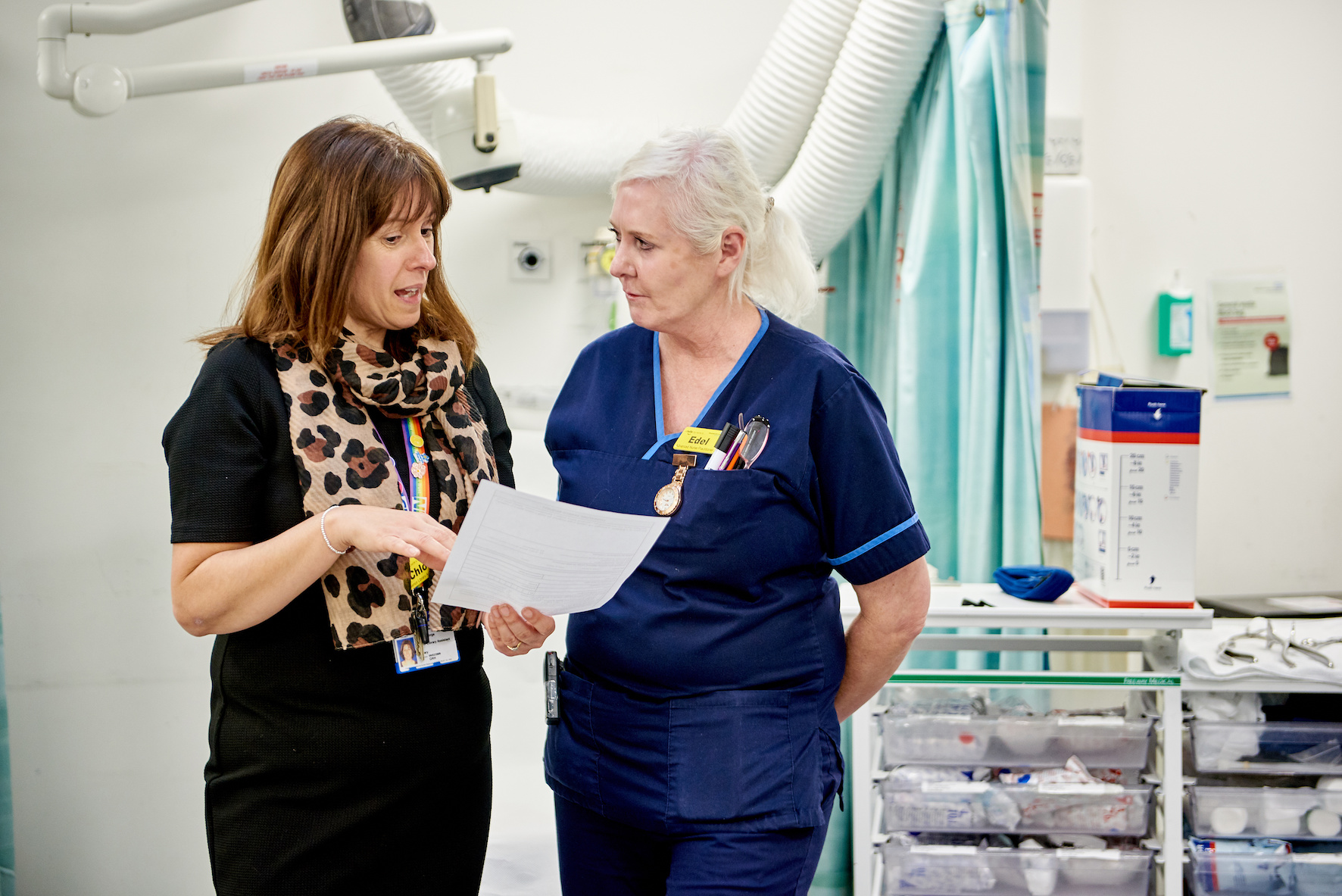 An image of 2 people standing in a ward discussing some patient information. On the left is a female medical professional with brown hair holding a paper and on the right is an older female medical professional with blonde hair listening to her.