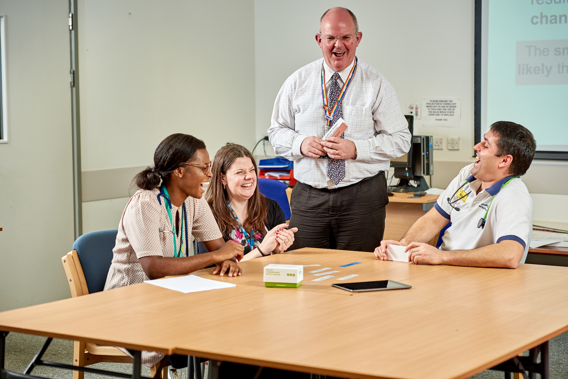 An image of a male standing up talking and laughing with 3 colleagues who are seated around a table.